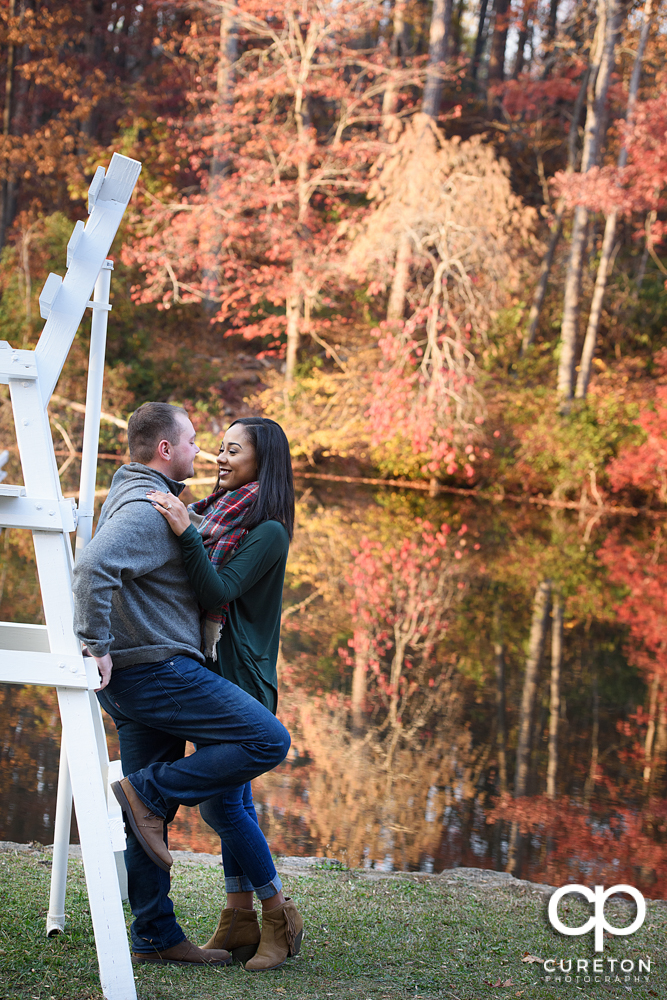 Engaged couple by the lake with fall leaves in the background.