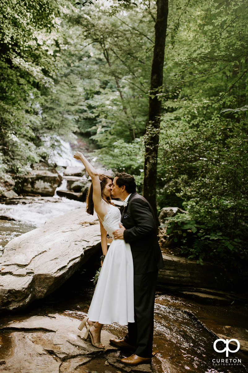 Bride holding her hand in the air while kissing her groom on a rock in the forest after their wedding at Pearson's Falls in Saluda,NC.