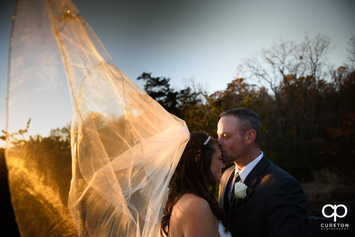 Bride and groom kissing as her veil blows in the wind and warm sunlight.