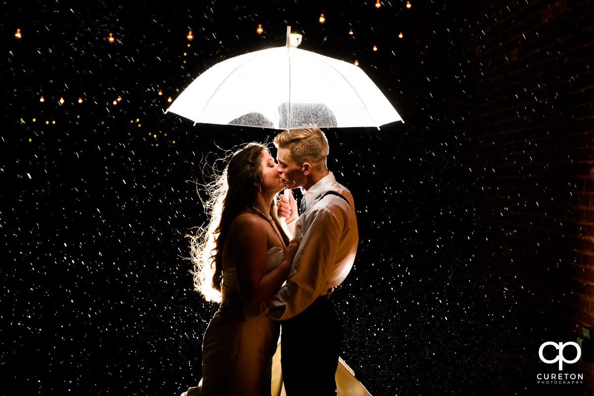 Bride and groom holding an umbrella kissing in the rain on the deck of The Old Cigar Warehouse.