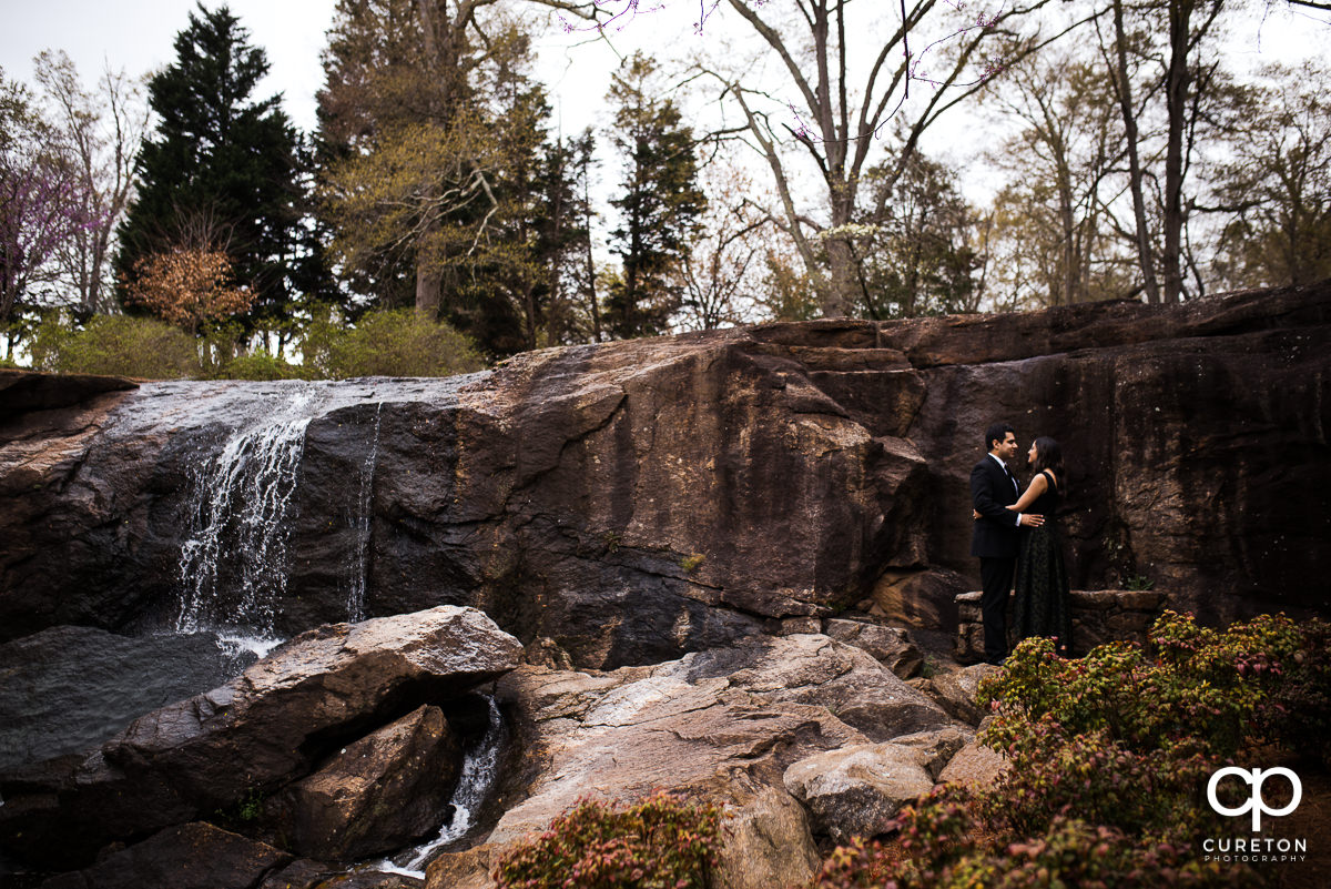 Future bride and groom hugging in front of a waterfall at their engagement session at The Rock Quarry Garden and Cleveland Park in Greenville,SC.