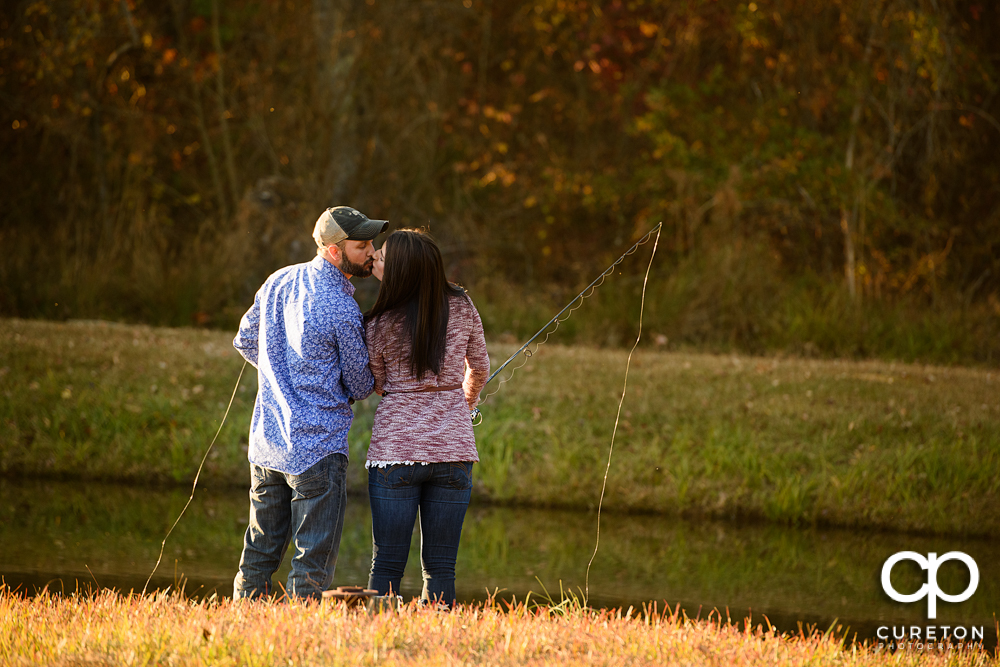 Engaged couple kissing while holding fishing poles.