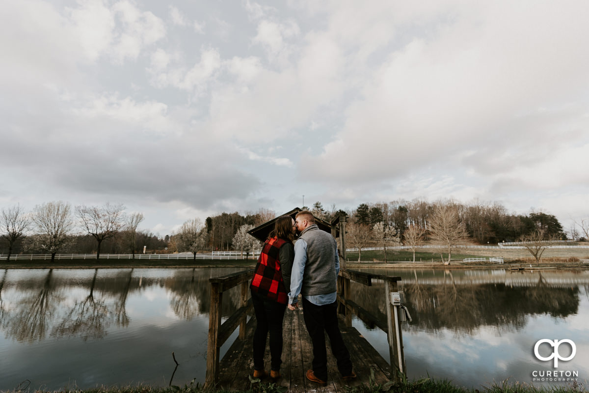 Engaged couple kissing on the dock in front of the pond during a pre wedding South Wind Ranch engagement session in Travelers Rest,SC.