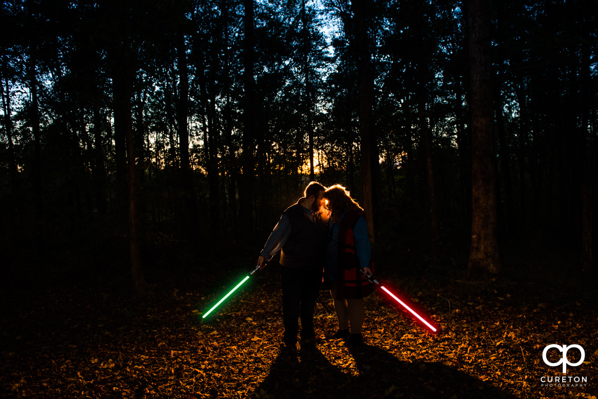 Future bride and groom holding lightsabers in the forest at their Star Wars themed engagement session.