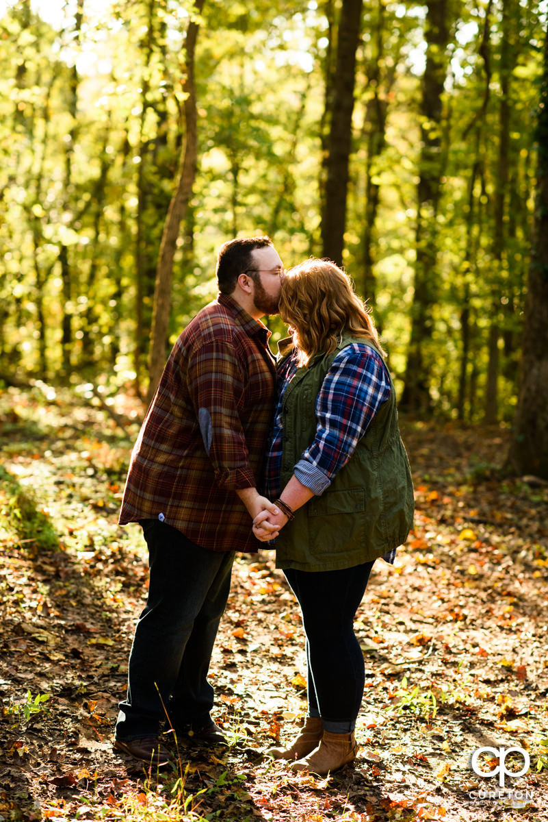 Groom kissing his bride on the forehead in the forest at their engagement session.