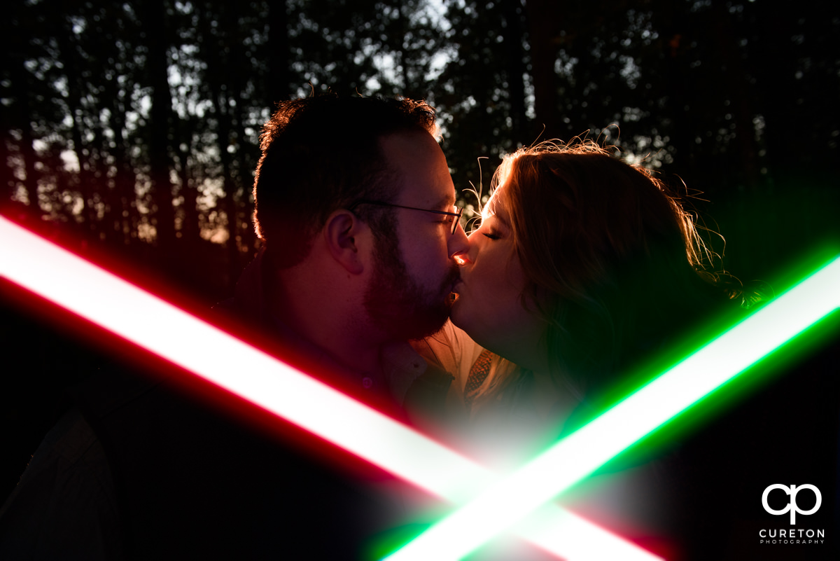 Engaged couple kissing behind a red and green lightsaber.