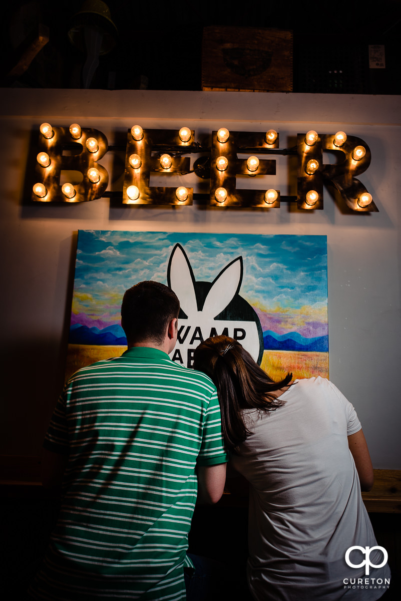 Woman laying her head on her fiancees shoulder as they sit in front of a beer sign during their Swamp Rabbit Brewery engagement session.