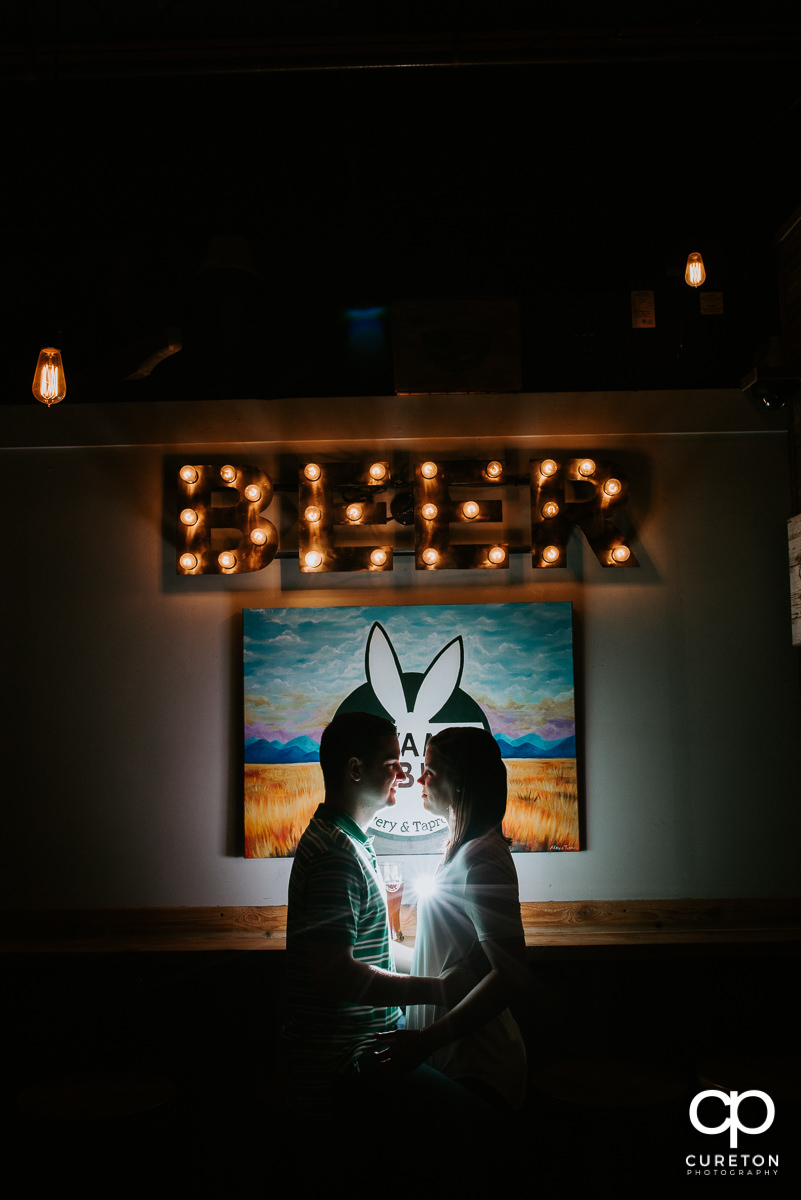 Future bride and groom hugging while having some beers in front of a lit beer sign during their Swamp Rabbit Brewery engagement session.