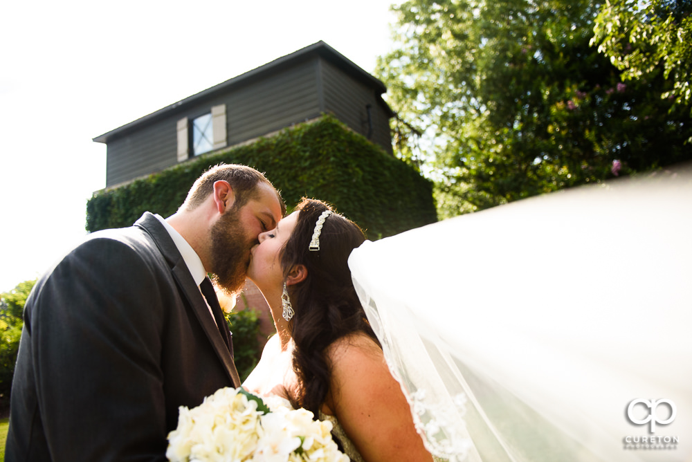 Bride and groom kissing withe her veil blowing in the wind after their wedding at The Loom a venue in Simpsonville, SC.