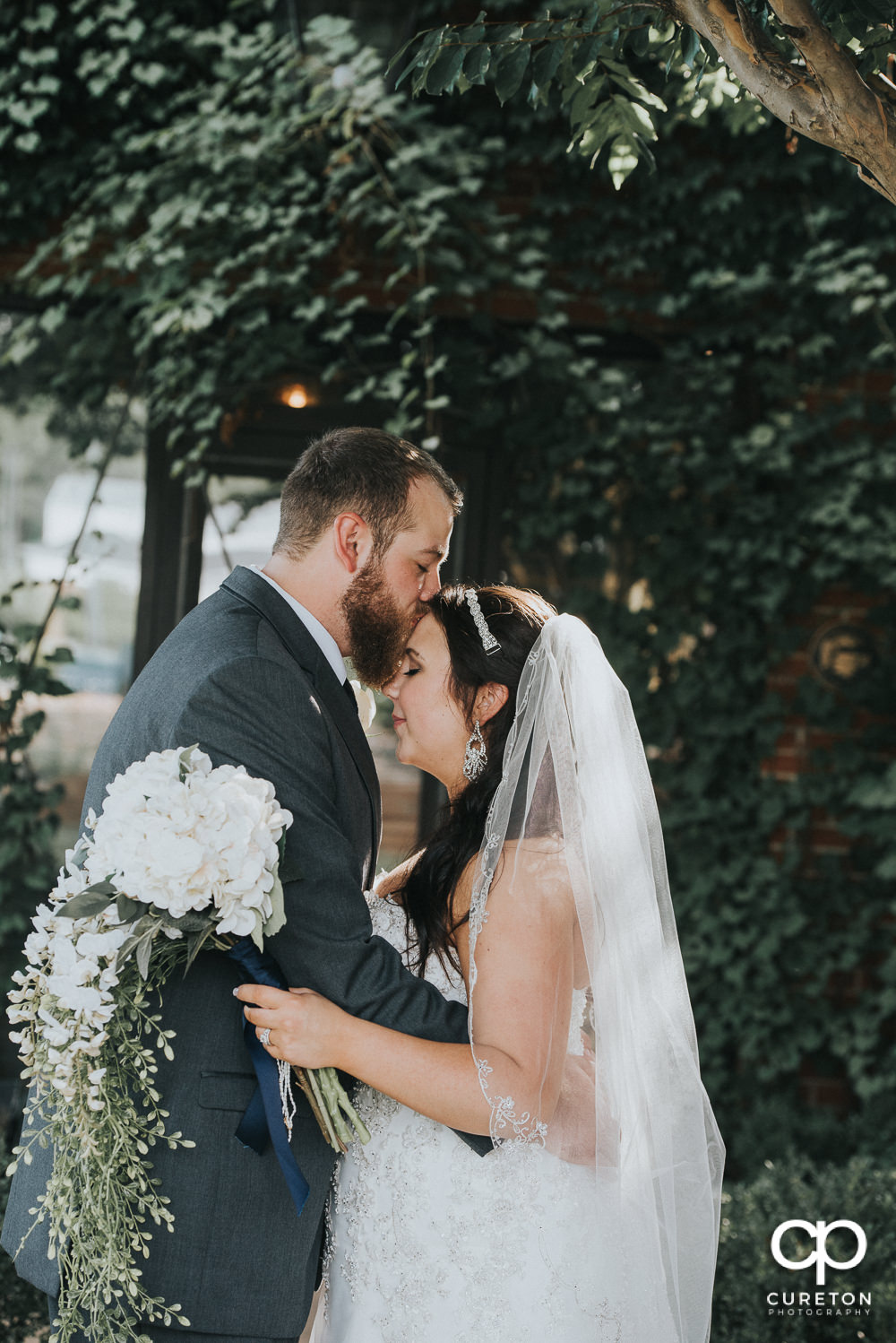 Groom kissing his bride on the forehead in front of a wall of ivy at The Loom after their wedding.