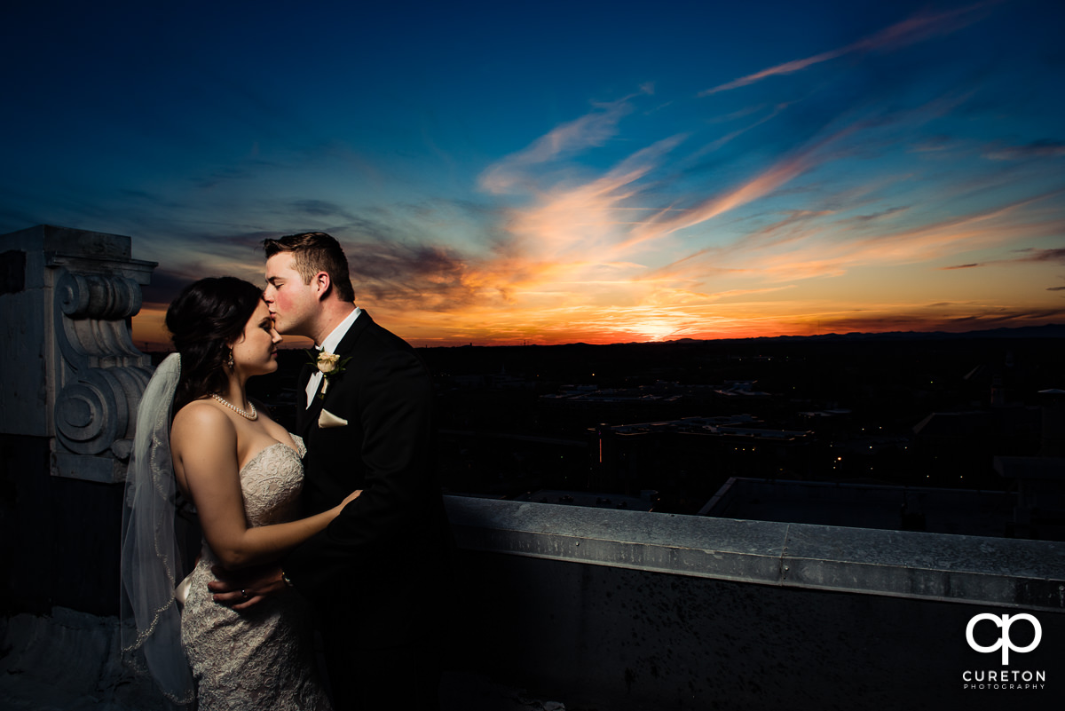 Groom kissing his bride on the forehead while watching an epic sunset over the Greenville SC skyline at their Westin Poinsett Hotel Wedding.