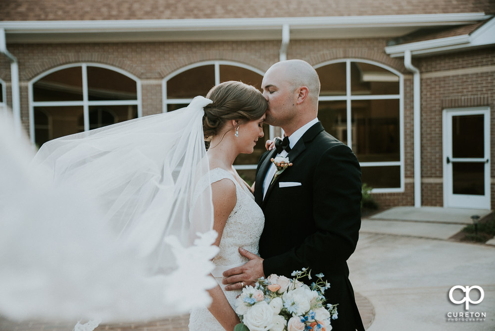 Groom kissing his bride on the forehead as her long veil blows in the wind.